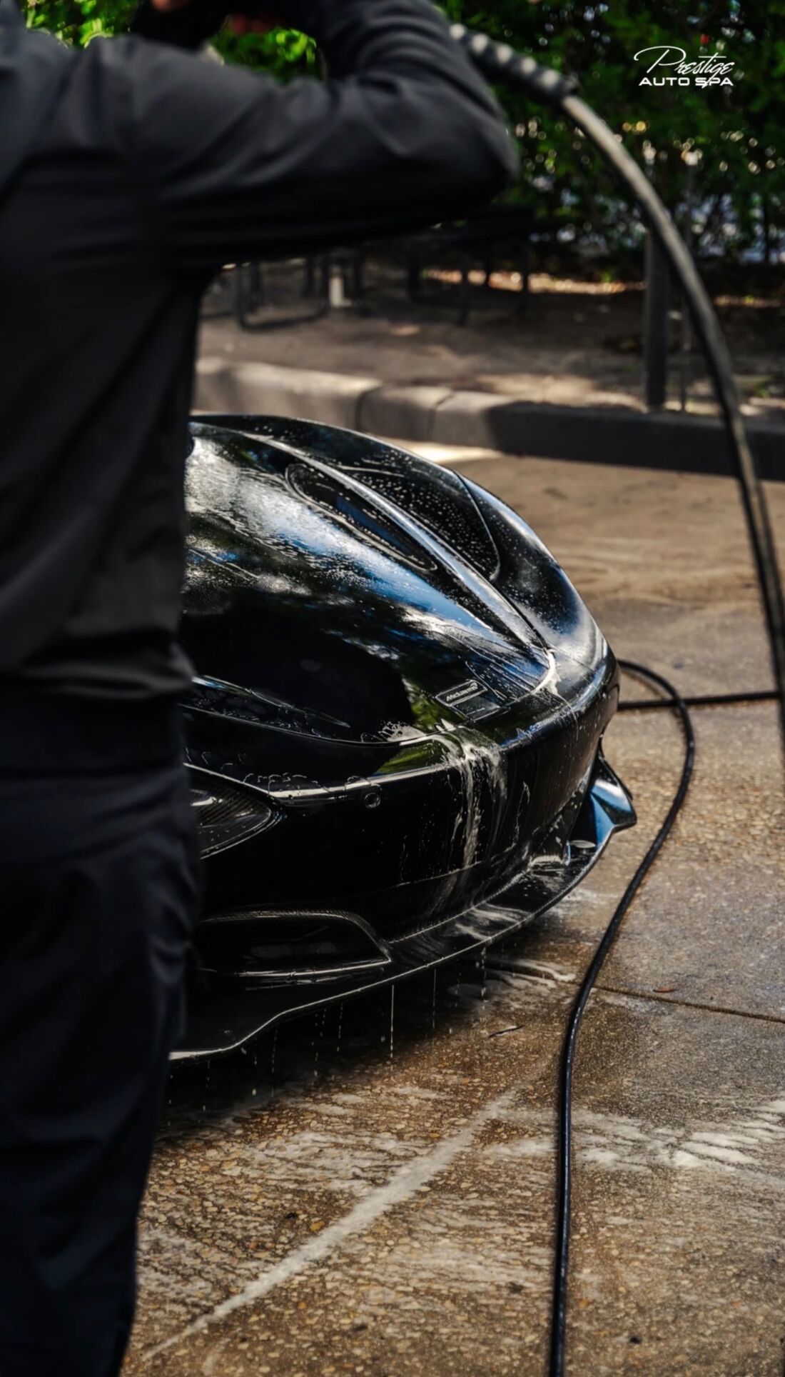 Black McLaren front end being rinsed while a detailer holds the pressure washer hose
